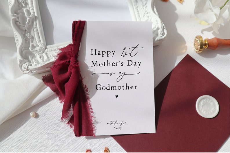 May include: A white card with a red ribbon tied around it. The card says "Happy 1st Mother's Day to my Godmother" with a small heart at the bottom. The card also says "with love from Avery".