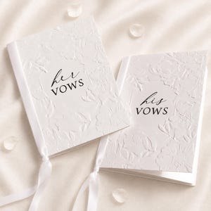 May include: Two white vow booklets featuring an embossed floral pattern on the covers. One booklet has the text "her VOWS" printed in black, and the other has "his VOWS" printed in black. Each booklet is tied with a white satin ribbon at the bottom.