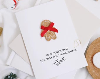 May include: A white Christmas card with a gingerbread man and a red bow. The card reads "Happy Christmas to a very special daughter" with the name "Layla" in cursive. The card is surrounded by other Christmas decorations.