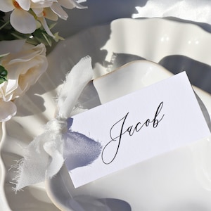 May include: A white place setting with a decorative edge, featuring a place card that reads "Jacob" in elegant black script. A white ribbon and floral arrangement add to the aesthetic. The scene is bathed in soft light.