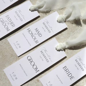 May include: White place cards with black text that reads "This seat is reserved for the" followed by the title of the person the seat is reserved for. The titles include "Maid of Honour", "Groom", and "Bride".