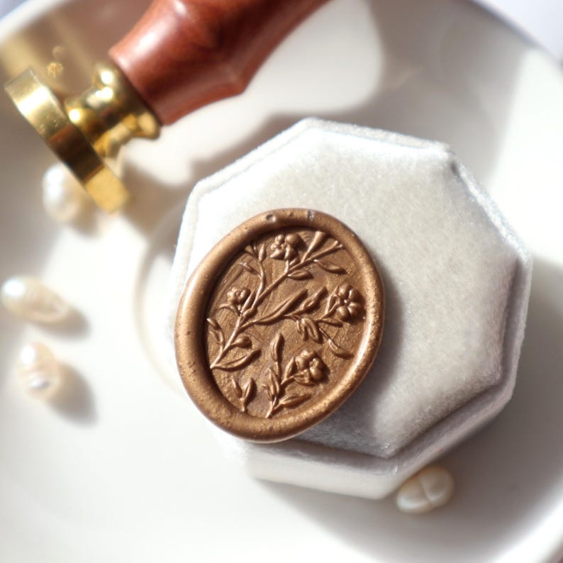 May include: A gold wax seal with a floral design. The seal is on a white velvet box.