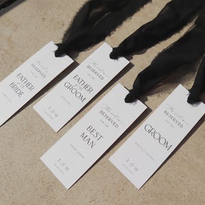 May include: Four white place cards with black calligraphy that reads "The next chapter" and "Reserved for the" followed by the title "Father of the Bride", "Father of the Groom", "Best Man", and "Groom". Each card has the name and date of the event printed below the title.