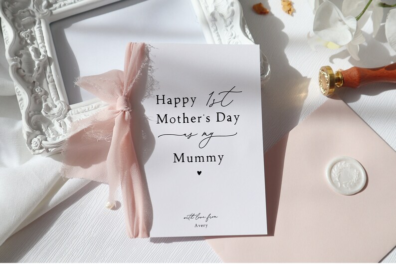 May include: A white card with a pink ribbon tied around it. The card says "Happy 1st Mother's Day as my Mummy" with a small heart at the bottom. The card also says "with love from Avery" at the bottom.