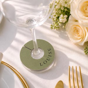 Op de afbeelding: Een ronde, saliegroene wijnglasmarker met een gleuf voor bevestiging aan de steel. De marker is bedrukt met de naam LOUISE in een donker, schreefloos lettertype. Het is geplaatst rond de voet van een heldere glazen steel op een wit kleed.
