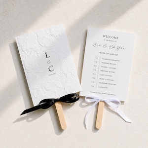 May include: Two white wedding programme fans with wooden handles. One features embossed floral patterns, the initials L & C, and the date 25.07.2017 with a black ribbon. The other displays a printed order of service and a white ribbon.