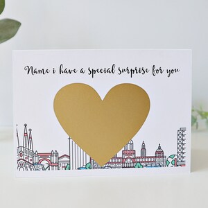 May include: A white card with a gold heart shape in the centre. The card says "Name, I have a special surprise for you". The heart shape is a scratch-off sticker. The card has a cityscape illustration at the bottom.