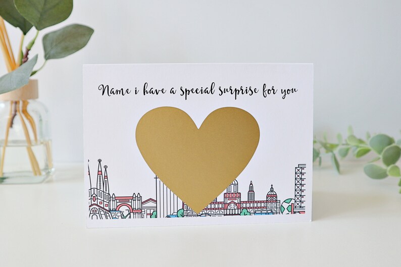 May include: A white card with a gold heart shape in the centre. The card says "Name, I have a special surprise for you". The heart shape is a scratch-off sticker. The card has a cityscape illustration at the bottom.