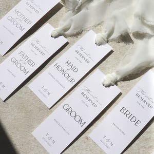 May include: Five white rectangular place cards with black text. Each card reads "This seat is reserved for the" followed by a title, such as "Mother of the Groom", "Father of the Groom", "Maid of Honour", "Groom", and "Bride". Each card also includes the name of the person the seat is reserved for and the date "T & M 24.05.2027".