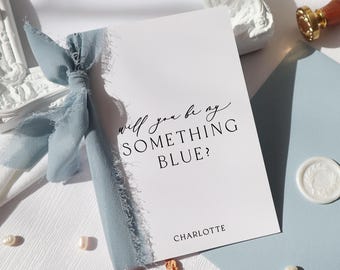 Peut inclure: Une carte rectangulaire blanche avec le texte noir Will you be my SOMETHING BLUE? et CHARLOTTE. La carte est ornée d'un ruban de soie effiloché bleu poussiéreux noué sur le côté gauche, accompagnée d'une enveloppe bleue assortie et d'un sceau de cire.