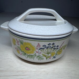 Op de afbeelding: Een keramische ovenschaal met deksel. De schaal is gebroken wit met een gespikkeld patroon en een bloemenontwerp met gele en blauwe bloemen. Het heeft een handvat op het deksel en twee zijhandvatten.