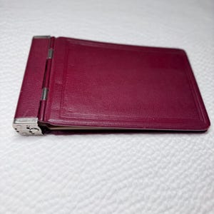 May include: A rectangular, deep magenta-colored wallet or card holder featuring a textured leather-like surface. The item includes a metallic silver-toned hinge mechanism on the left side with visible circular rivets and a smooth, clean edge finish.