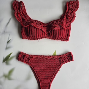Puede incluir: Un conjunto de bikini de ganchillo de dos piezas en color rojo intenso. La parte superior presenta una textura acanalada, un escote redondo y tirantes con volantes y detalles intrincados de encaje. La braga a juego tiene un corte de pierna alto.