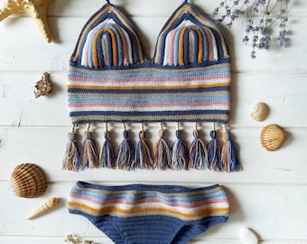 Op de afbeelding: Een tweedelige gehaakte bikiniset bestaande uit een haltertop en een bijpassend broekje. Het ontwerp bevat horizontale strepen in tinten blauw, wit, roze en mosterdgeel. De top is afgewerkt met een franjezoom en kleine houten kralen.
