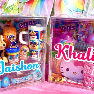 May include: Two clear plastic boxes filled with toys and treats. One box features a Chase the Paw Patrol plush, a mug, and a container with the name "Jaishon". The other box has a Hello Kitty theme with the name "Khali".