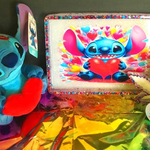 Peut inclure: Un personnage Stitch bleu et blanc tenant un cœur rouge, entouré de cœurs roses, jaunes et bleus. Le personnage est sur un plateau roulant blanc avec une bordure de paillettes. Il y a une feuille de marijuana en verre transparent et un petit plat rond avec le même personnage Stitch.