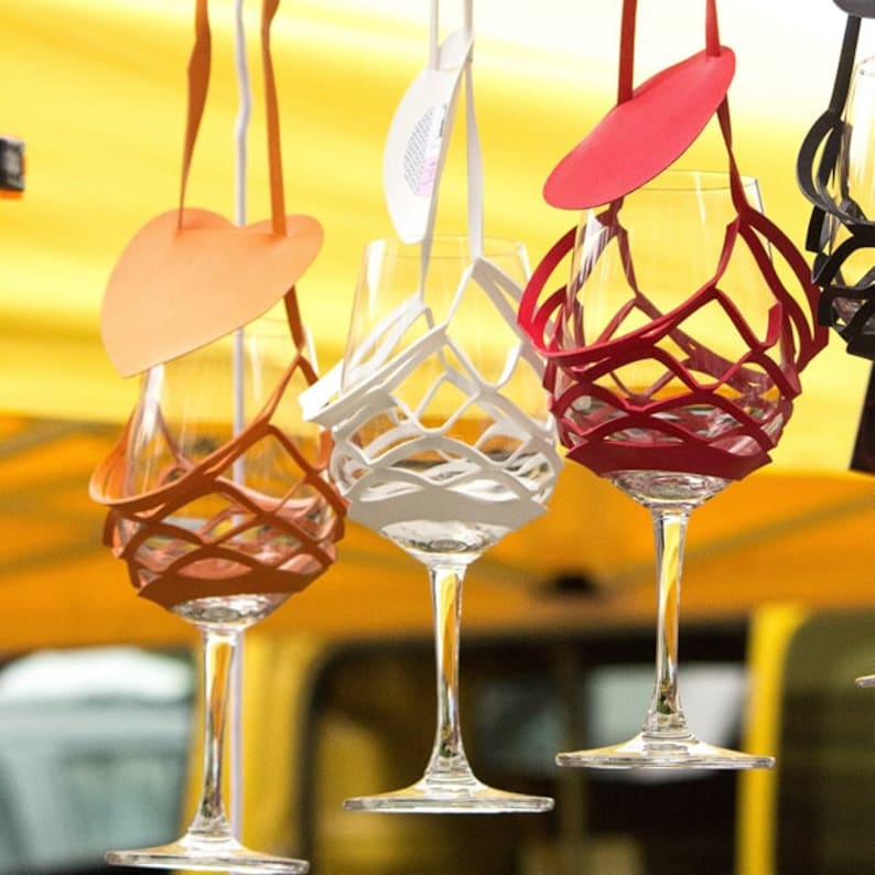 May include: Four wine glasses with colorful, decorative, fabric holders. The holders are attached to the glasses with string and are shaped like hearts and circles. The glasses are hanging from the ceiling.