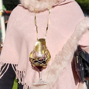 May include: A gold-colored metal wine glass holder with a glass of red wine inside. The holder is attached to a gold chain and worn around the neck.