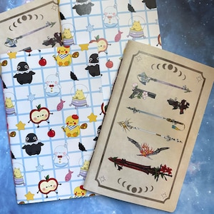 May include: Notebooks with whimsical illustrations. One notebook features a repeating pattern of cartoon characters, apples, and stars on a blue grid. Another has weapon illustrations. The covers are light brown and white.