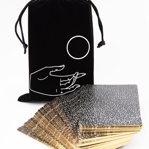 May include: A black velvet bag with a white hand and circle design, holding a deck of black and gold tarot cards with white speckles.