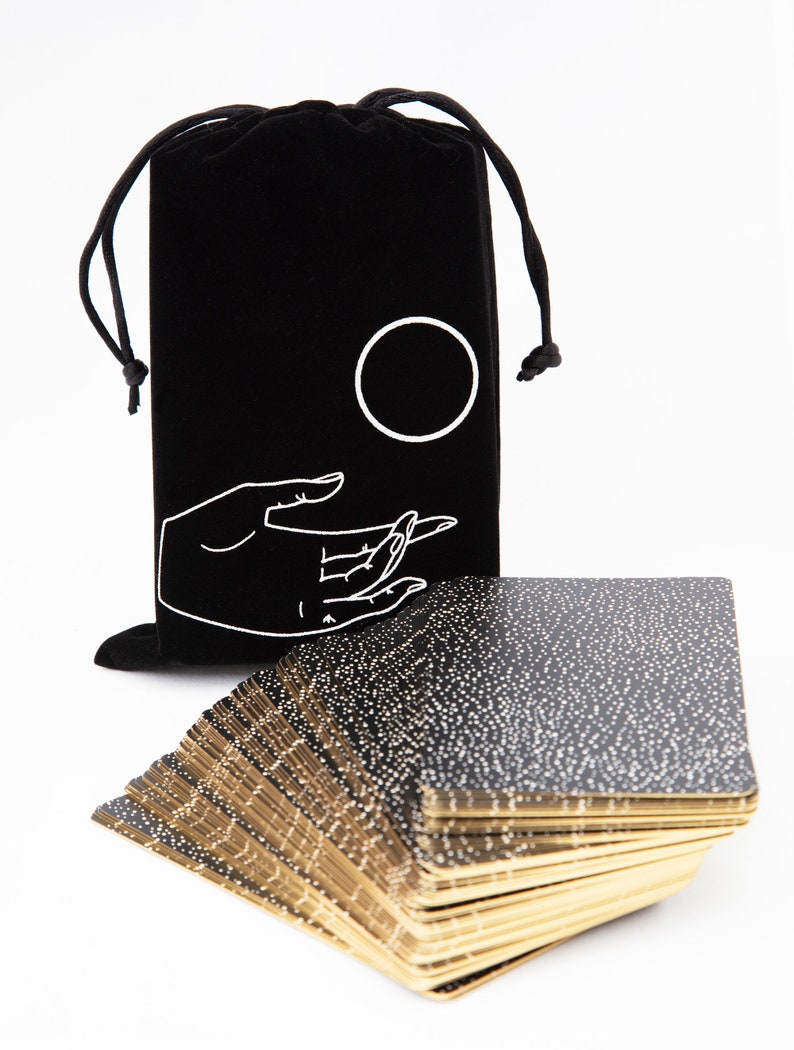 May include: A black velvet bag with a white hand and circle design, holding a deck of black and gold tarot cards with white speckles.