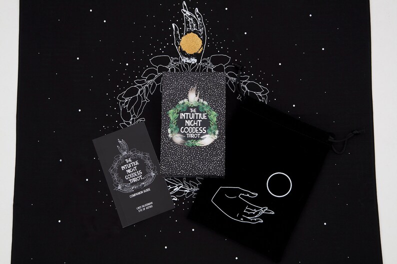 May include: A black velvet bag with a white hand and a circle design. The bag is next to two black and white cards with the text "The Intuitive Night Goddess Tarot" on them. The cards are on a black background with white stars.