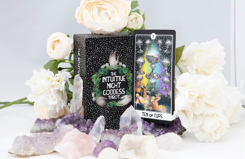 Pu&ograve; includere: Una scatola nera di mazzo di tarocchi con lettere dorate che recitano "The Intuitive Night Goddess Tarot". La scatola &egrave; circondata da cristalli bianchi e viola e fiori bianchi. Una carta dei tarocchi con un'immagine colorata di dieci coppe &egrave; visibile sullo sfondo.