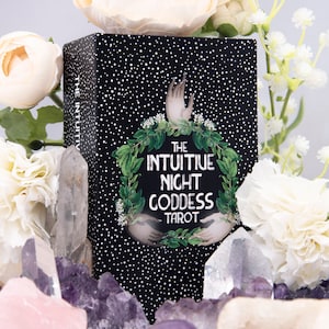 Pu&ograve; includere: Una scatola nera di mazzo di tarocchi con pois bianchi e il testo "The Intuitive Night Goddess Tarot". La scatola &egrave; circondata da fiori bianchi e rosa e cristalli di quarzo trasparenti.