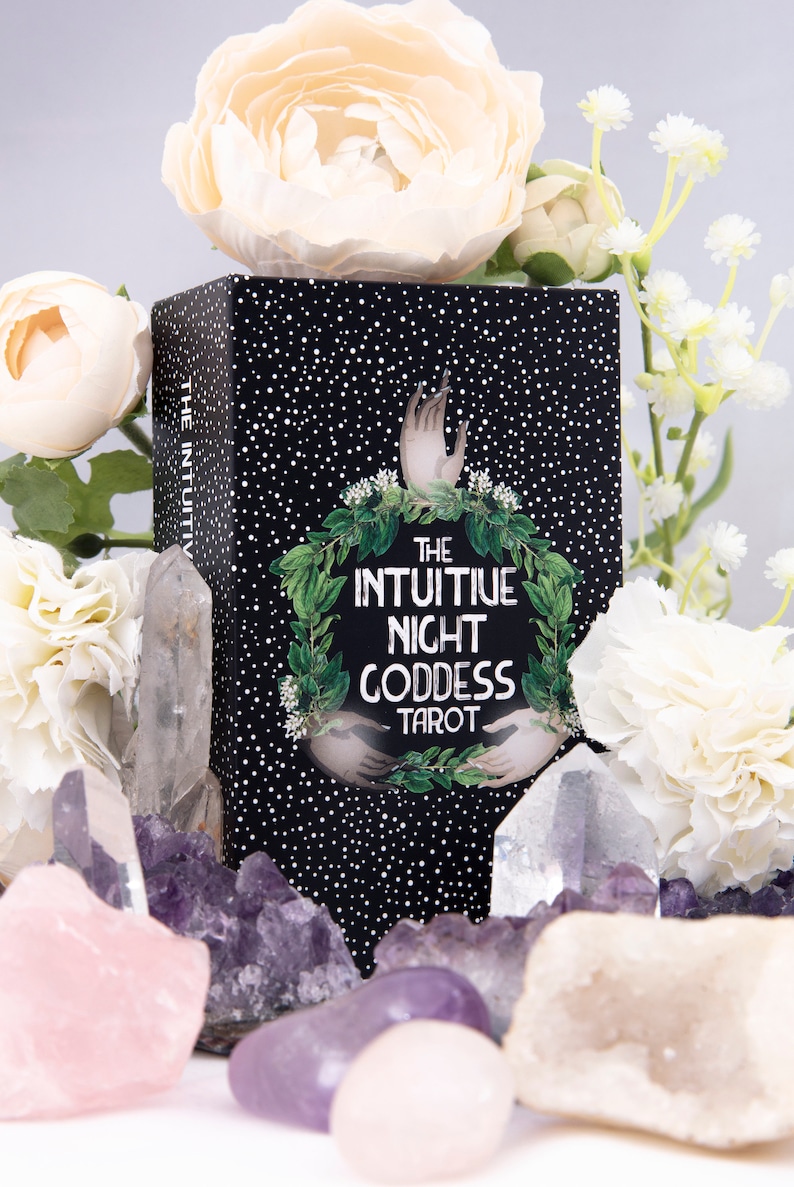 Pu&ograve; includere: Una scatola nera di mazzo di tarocchi con pois bianchi e il testo "The Intuitive Night Goddess Tarot". La scatola &egrave; circondata da fiori bianchi e rosa e cristalli di quarzo trasparenti.