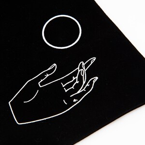 May include: A black velvet pouch with a white outline of a hand reaching up to a circle.