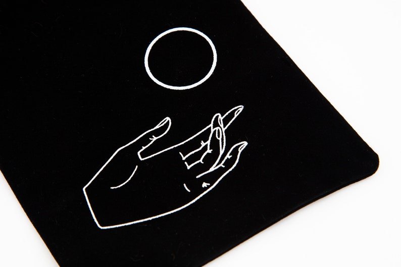 May include: A black velvet pouch with a white outline of a hand reaching up to a circle.