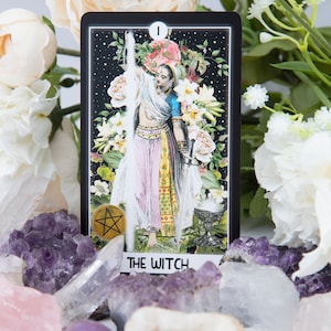 Pu&ograve; includere: Una carta dei tarocchi con sfondo nero e una donna in un abito bianco che tiene una spada. La carta &egrave; circondata da fiori bianchi e cristalli. La carta recita "La Strega" e ha un simbolo di pentagramma nell'angolo in basso a sinistra.