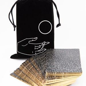 Pu&ograve; includere: Un mazzo di carte da tarocchi nere e dorate con macchie bianche sul retro, impilate in un mucchio. Le carte sono in una borsa di velluto nero con una mano bianca e un disegno circolare.