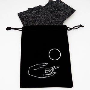 May include: Black velvet bag with a white drawstring closure. The bag has a white design of a hand holding a circle. The bag is filled with a deck of black cards with white specks.