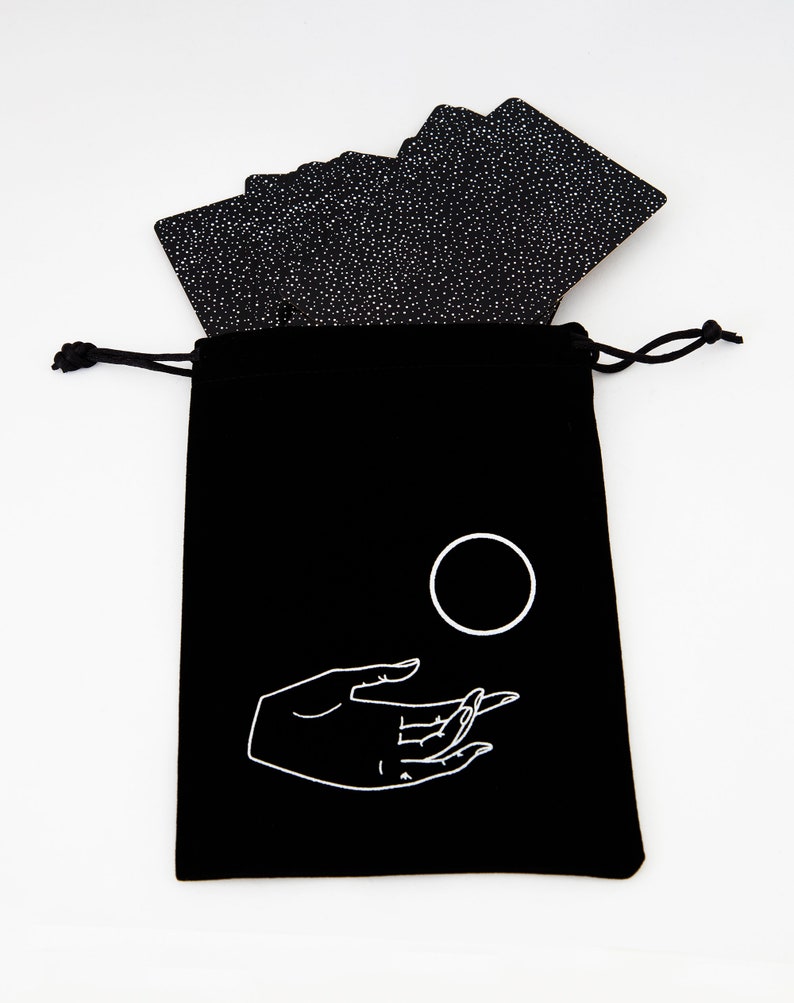 May include: Black velvet bag with a white drawstring closure. The bag has a white design of a hand holding a circle. The bag is filled with a deck of black cards with white specks.