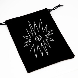 May include: Black drawstring bag with a white graphic of a stylized eye within a starburst design. The bag is made of a soft material and has a drawstring closure. The bag is on a white background.
