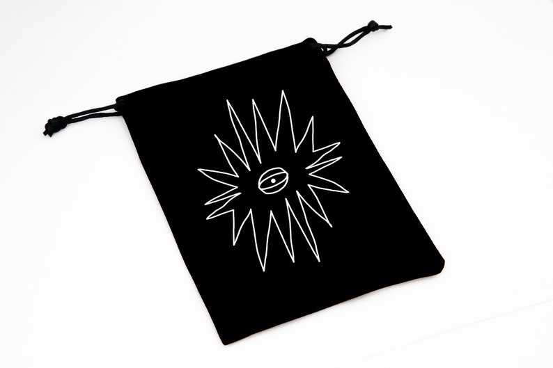 May include: Black drawstring bag with a white graphic of a stylized eye within a starburst design. The bag is made of a soft material and has a drawstring closure. The bag is on a white background.