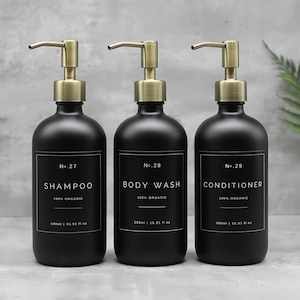 Peut inclure: Trois flacons en verre noir avec des pompes dorées et des étiquettes blanches. Les flacons sont étiquetés "No. 27 Shampooing 100% Bio", "No. 28 Gel Douche 100% Bio" et "No. 29 Après-Shampoing 100% Bio". Chaque flacon contient 500 ml (16,91 fl oz).