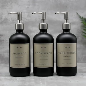Könnte beinhalten: Drei schwarze Pumpflaschen mit silbernen Verschlüssen und Etiketten. Die Flaschen sind mit "No. 27 Shampoo 100% Bio", "No. 28 Duschgel 100% Bio" und "No. 29 Conditioner 100% Bio" beschriftet.