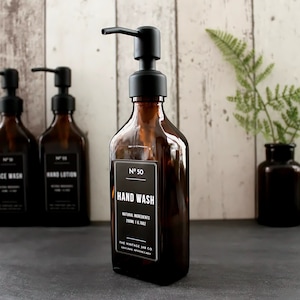 Puede incluir: Tres botellas de vidrio ámbar con tapas de bomba negras y etiquetas. Las botellas están etiquetadas como "Face Wash", "Hand Lotion" y "Hand Wash". Las botellas están sobre una superficie negra.