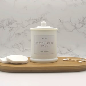 Może przedstawiać: Biały ceramiczny słoik na świecę z pokrywką i etykietą z napisem "No. 35 Cotton Wool Pads 100% Organic". Słoik na świecę stoi na drewnianej tacy z wacikami kosmetycznymi i patyczkami kosmetycznymi.