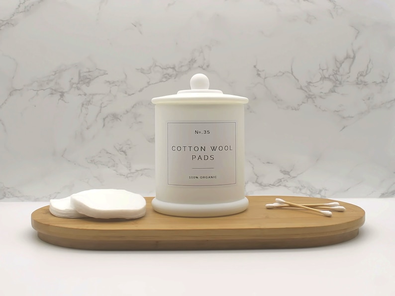 Może przedstawiać: Biały ceramiczny słoik na świecę z pokrywką i etykietą z napisem "No. 35 Cotton Wool Pads 100% Organic". Słoik na świecę stoi na drewnianej tacy z wacikami kosmetycznymi i patyczkami kosmetycznymi.