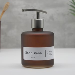 May include: A frosted amber glass soap dispenser with a silver-toned metal pump. The white rectangular label features the black text Hand Wash, 250ml, and a list of three bullet points: refill, reuse, and repeat, separated by a vertical line.