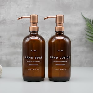 Op de afbeelding: Twee amberkleurige glazen flessen met koperen pompen, met het label "No. 03 Handzeep Natuurlijke ingrediënten 500 ml | 16,91 fl oz" en "No. 04 Handlotion Natuurlijke ingrediënten 500 ml | 16,91 fl oz".