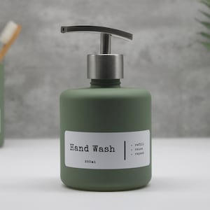 May include: A sage green hand wash dispenser with a silver pump. The label reads "Hand Wash" and "850ml". The dispenser is on a white surface, with a green toothbrush holder and toothbrushes in the background.