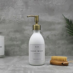 May include: A white hand wash dispenser with a gold-colored pump, labeled "Hand Wash" and "100% Organic." A small wooden brush and a jar of cotton pads are also visible. The dispenser holds 300ml or 10.14 fl oz.