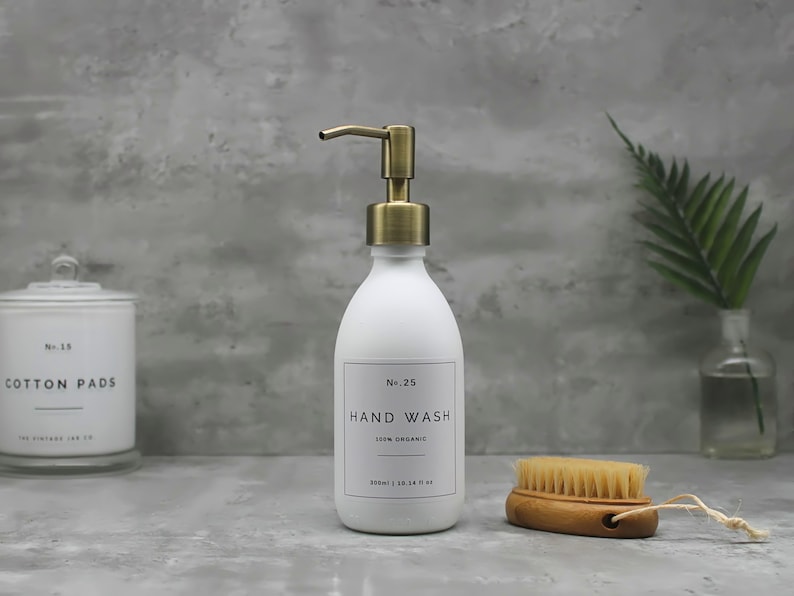 May include: A white hand wash dispenser with a gold-colored pump, labeled "Hand Wash" and "100% Organic." A small wooden brush and a jar of cotton pads are also visible. The dispenser holds 300ml or 10.14 fl oz.