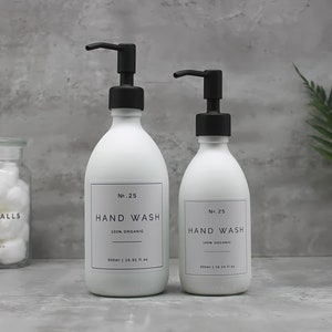 Könnte beinhalten: Zwei weiße Keramik-Handseifenflaschen mit schwarzen Pumpen und Etiketten mit der Aufschrift "No. 25 HAND WASH 100% ORGANIC" und "No. 25 HAND WASH 100% ORGANIC". Die größere Flasche ist 500 ml (16,91 fl oz) und die kleinere Flasche ist 300 ml (10,14 fl oz).