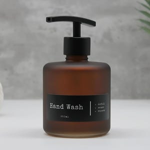 May include: A frosted amber glass bottle with a black pump dispenser. A black rectangular label on the front features the white text Hand Wash, 250ml, and a list of three items: refill, reuse, and repeat, separated by a vertical white line.