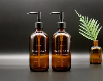 Op de afbeelding: Twee amberkleurige glazen flessen met zwarte pompjes en etiketten. De fles links heeft het opschrift "No. 02 DISH SOAP NATURAL INGREDIENTS 500ml | 16.9 fl oz". De fles rechts heeft het opschrift "No. 03 HAND SOAP NATURAL INGREDIENTS 500ml | 16.9 fl oz".